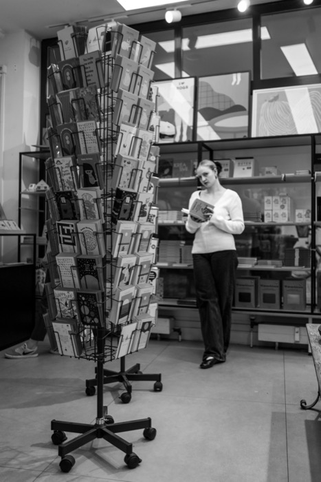 Woman reading a book in a stationery shop beside a tall rotating rack of greeting cards