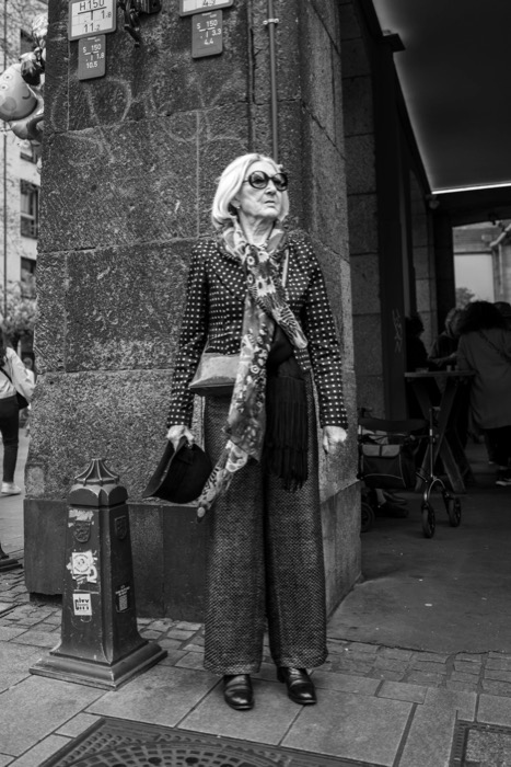 Older woman in sunglasses, polka-dot jacket and patterned scarf standing against a stone corner