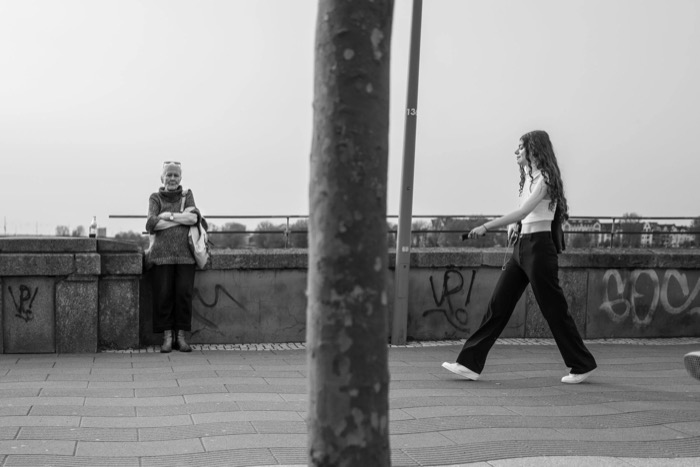 Young woman striding past a riverside wall while an older woman leans against it with arms folded, tree trunk splitting the frame