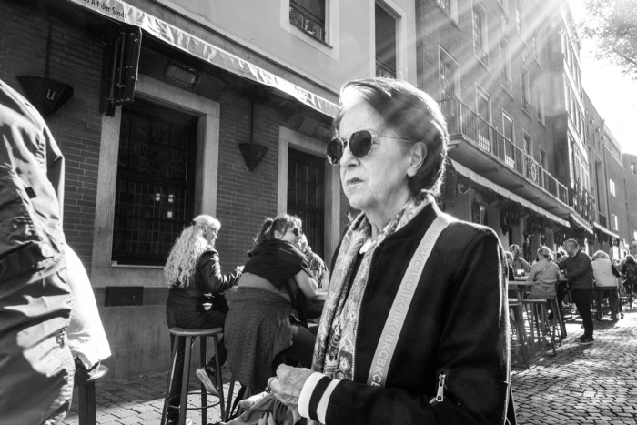 Woman in dark sunglasses and a track jacket walks past bar stools and drinkers on a sunlit cobbled street