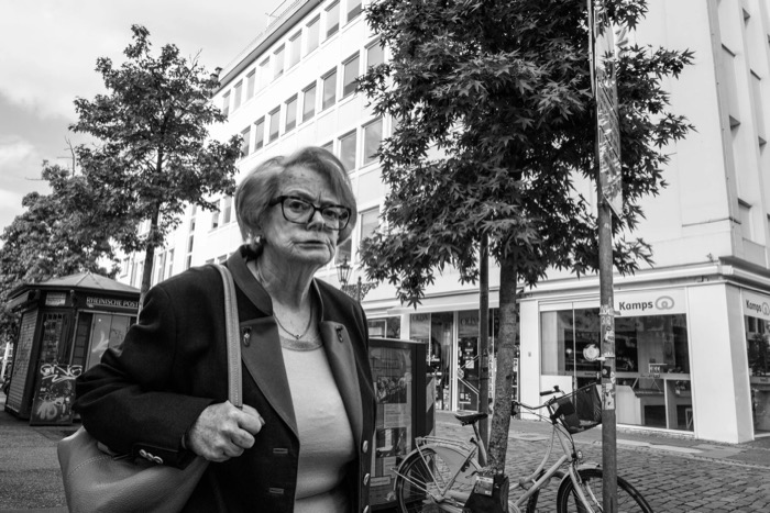 Elderly woman with glasses and a handbag pauses on a shopping street, bicycle and bakery behind