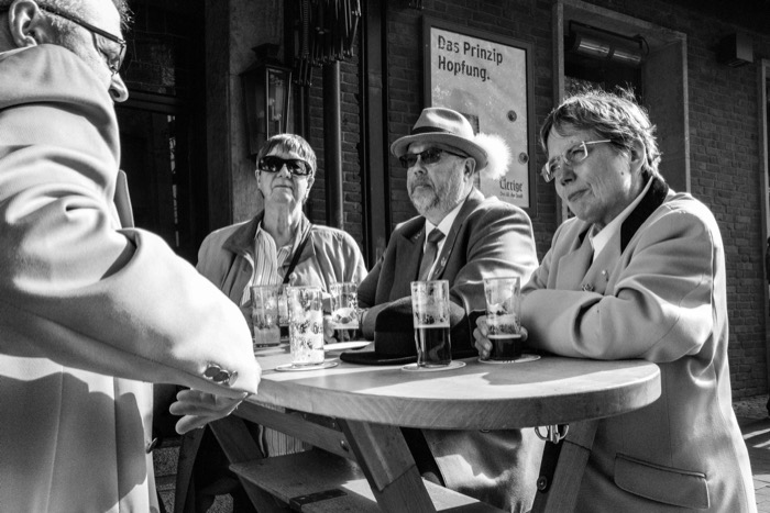 Three older people with beer glasses at an outdoor pub table, a waiter's arm in the foreground