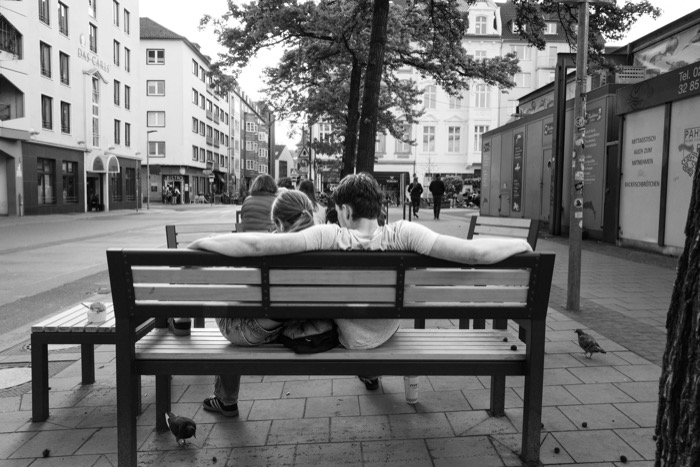 Two people on a pedestrian-zone bench seen from behind, arms along the backrest, pigeons on the pavement