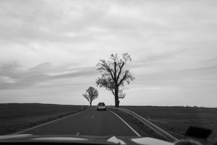 View through a windscreen of a car ahead on a straight country road beside two bare trees