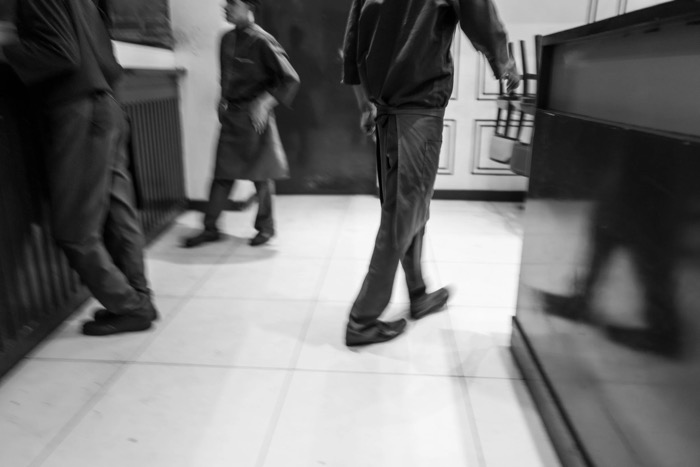 Blurred figures in aprons striding across a tiled floor, motion smearing the whole frame