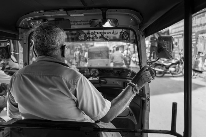 Auto-rickshaw driver seen from the back seat, hand on the throttle, busy market street through the windscreen