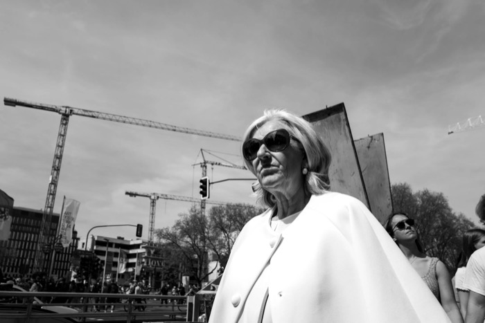 Older woman in a white cape and large sunglasses walking past a skyline of construction cranes