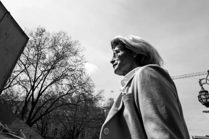 Low-angle profile of a white-haired woman in a coat, bare trees and a crane behind her