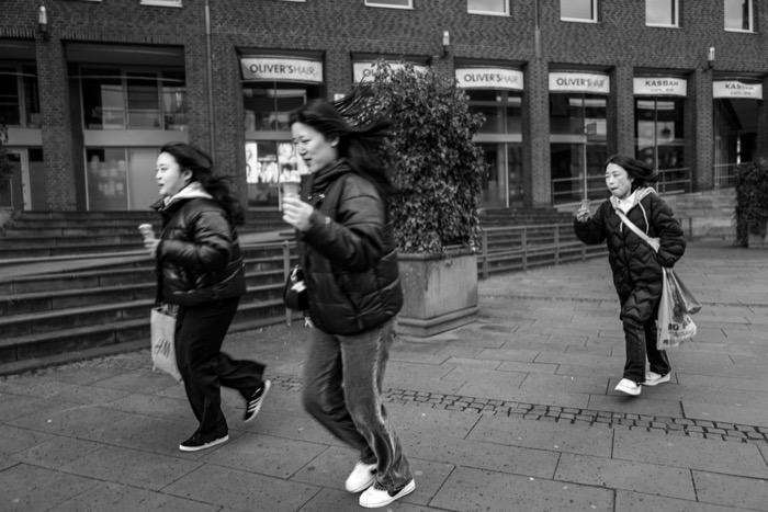 Three women in puffer jackets hurrying across a windy town square holding ice cream cones