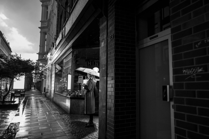Woman under an umbrella pausing at a shop window on a rain-slicked cobbled street