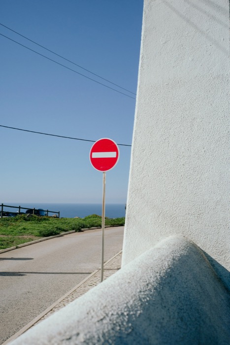 Red no-entry sign beside a whitewashed wall on a coastal road, blue sea on the horizon