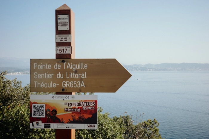 Wooden coastal trail signpost reading Pointe de l'Aiguille, Sentier du Littoral, above a hazy blue bay