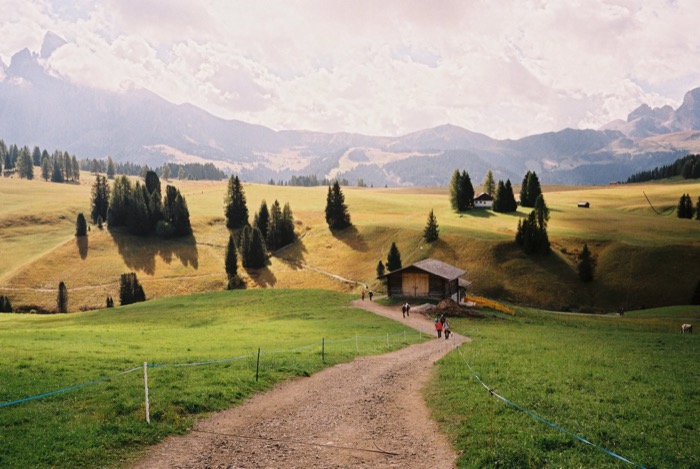 Gravel path curving through high alpine meadows past a wooden barn toward distant peaks
