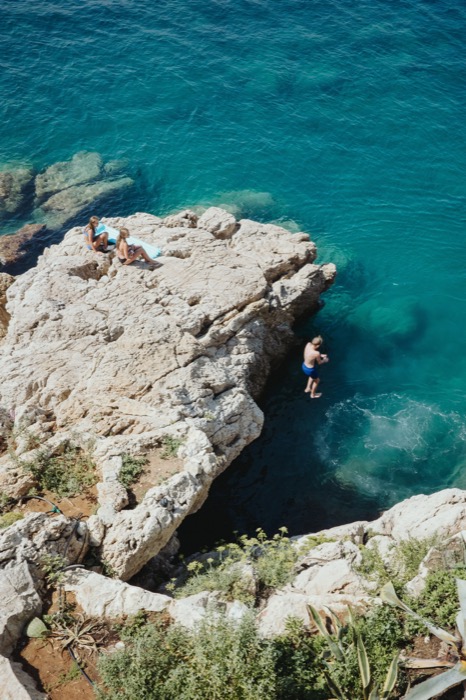 Swimmer stepping off a limestone ledge into turquoise sea, two sunbathers on the rocks above