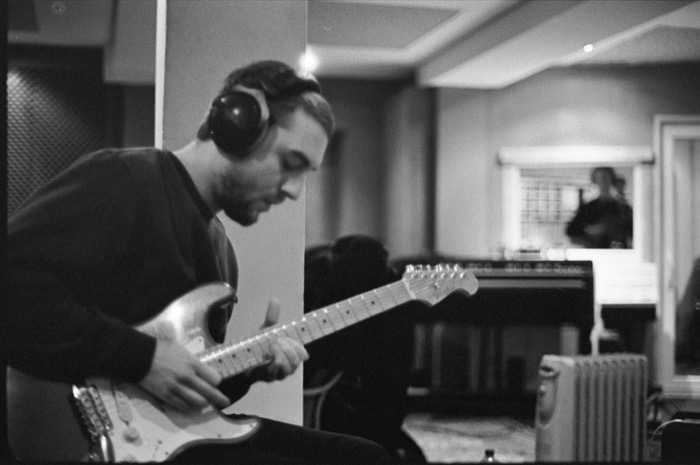 Guitarist in headphones bends over his instrument in a recording booth, engineer visible through the glass