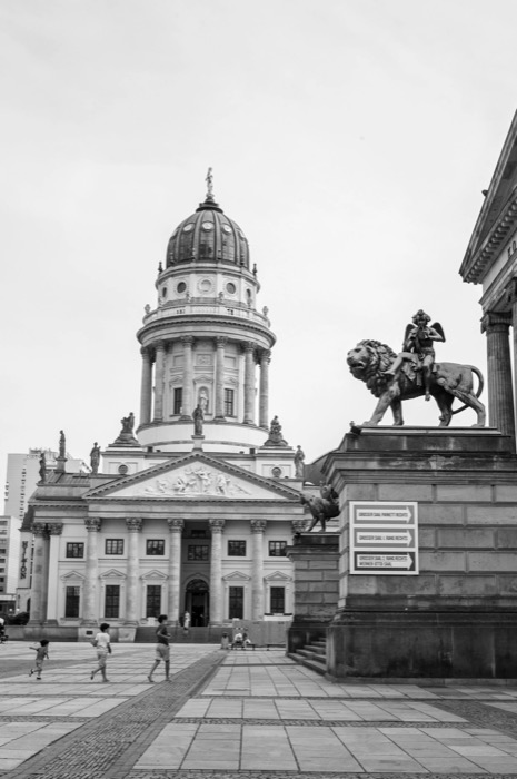 Children running across a wide square before a domed classical church, bronze lion-and-rider statue at right