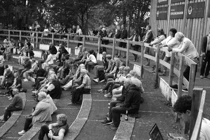 Crowd seated on curved concrete steps of an open-air amphitheatre, others leaning on a wooden rail