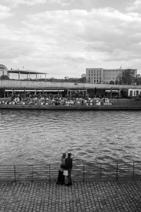 Couple standing close at a river railing on cobblestones, a crowded terrace on the far bank beyond the water