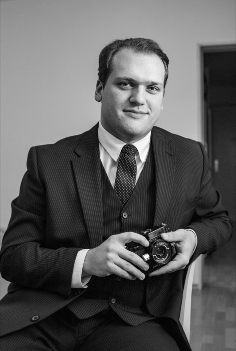 Young man in a three-piece pinstripe suit seated, holding a rangefinder camera in both hands