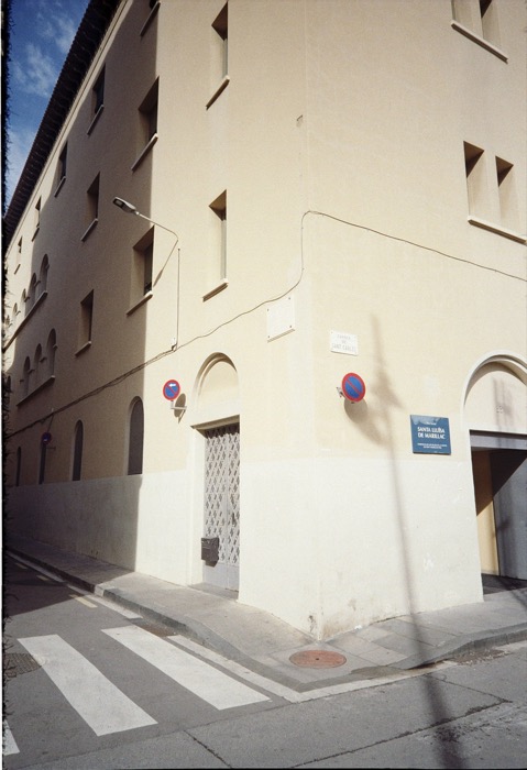 Sunlit corner of a plain cream apartment block with no-parking signs and a street plaque reading Sant Carles