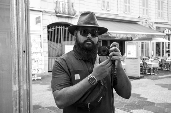 Bearded man in a wide-brimmed hat and sunglasses photographing his reflection in a shop window