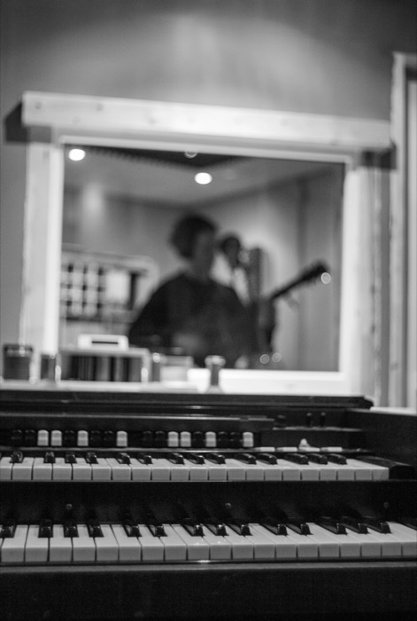 Organ keyboards in sharp focus, a guitarist blurred through a studio window beyond