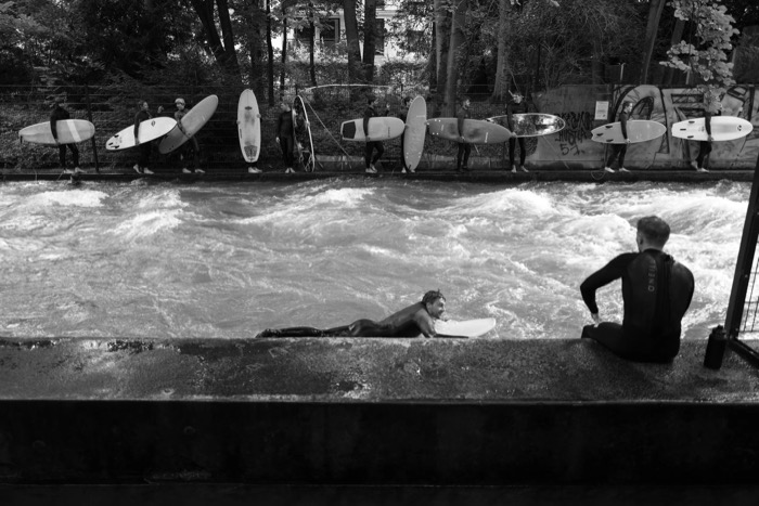 Surfers queued along a railing with their boards above a standing river wave in a city park