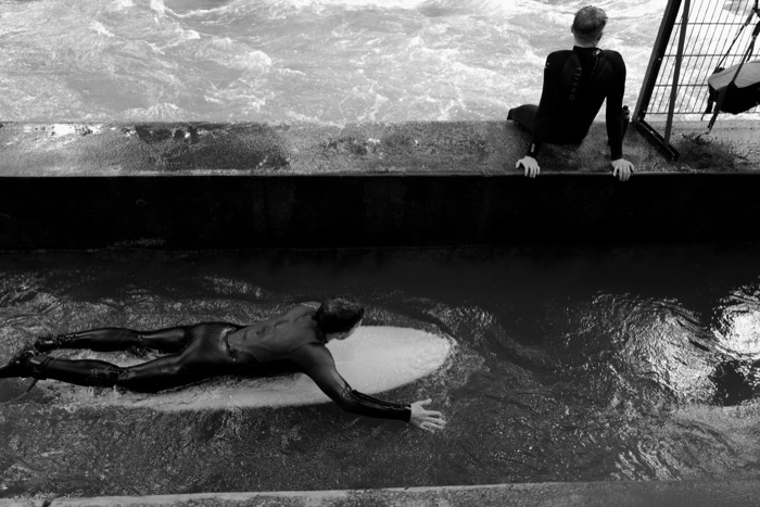 Surfer lying flat on a board in a channel of dark river water while another in a wetsuit sits on the wall above