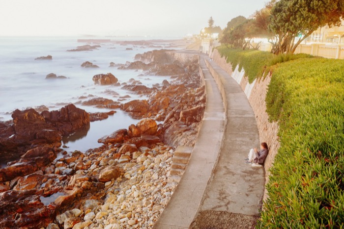 Woman sitting alone on a coastal promenade wall above rocks and long-exposure surf at dusk