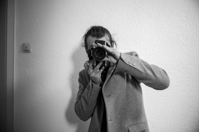 Person in a grey coat raises a film SLR to their face, hiding it, against a plain white wall