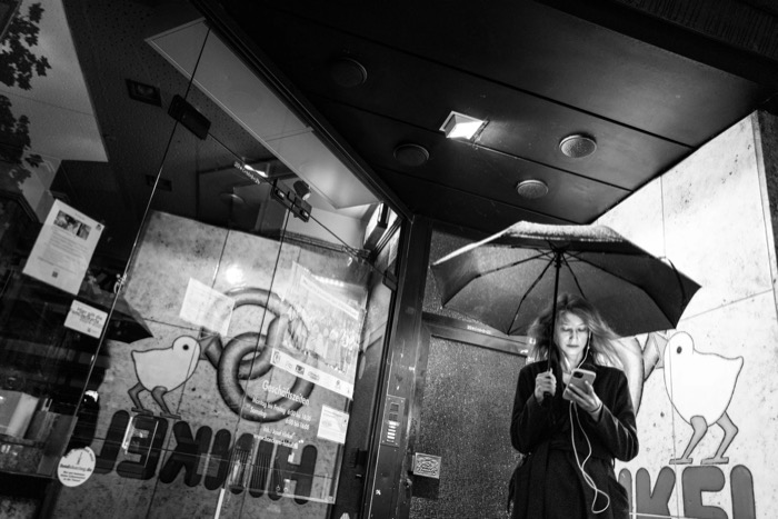 Woman under an umbrella checks her phone with earphones in, beside a shop window with a duck logo