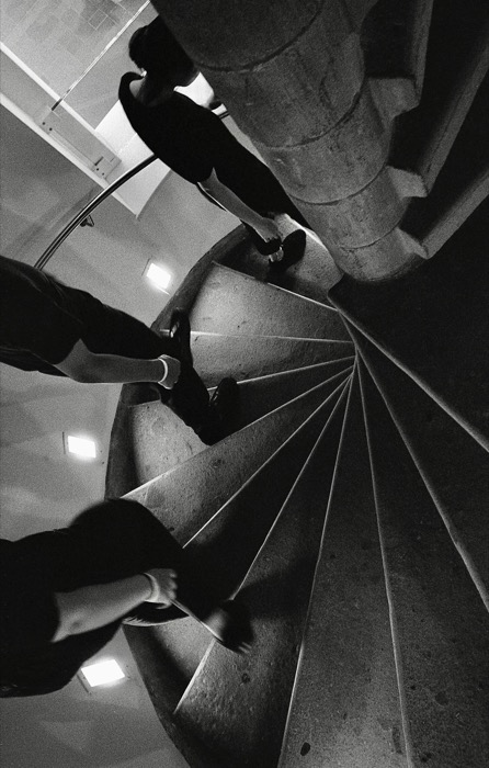 Looking straight down a concrete spiral staircase as three people descend, legs cut by hard shadow