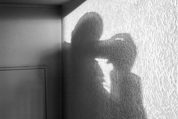 Shadow of a hat-wearing figure raising a camera, cast across a textured white interior wall