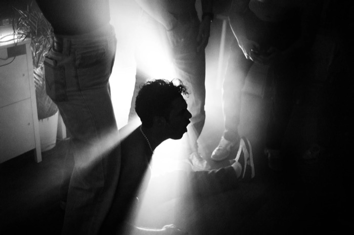 Man's head in silhouette against a blast of light at a crowded party, legs and sneakers all around