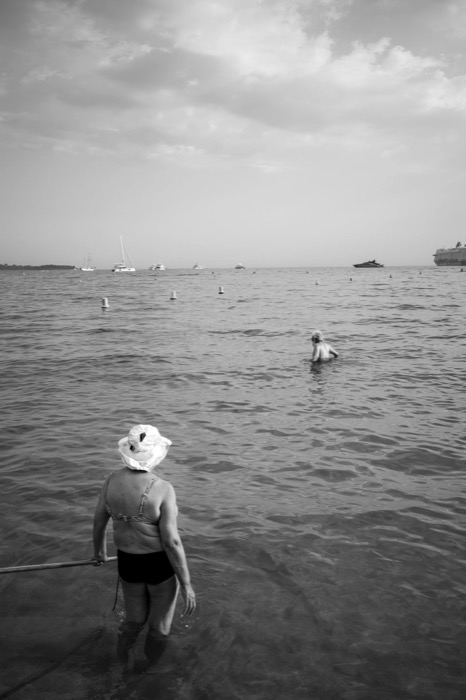 Woman in a floppy sun hat wading into the sea holding a rail, another bather and yachts beyond