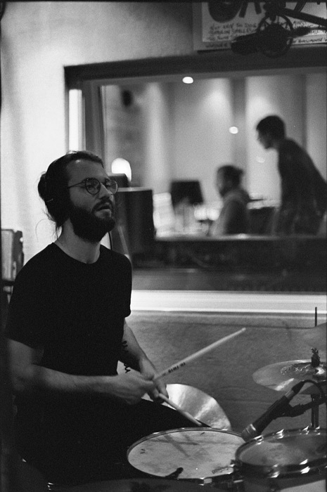 Bearded drummer in headphones and round glasses playing behind a control-room window