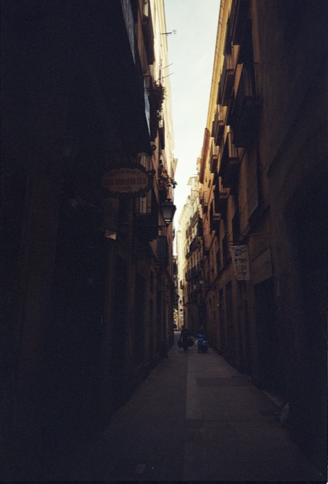 Narrow old-town alley in deep shadow, a lone figure with bags at the sunlit far end