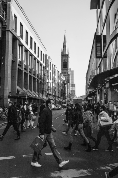 Shoppers crossing a city street, one man in a mask carrying a bag, church spire closing the view