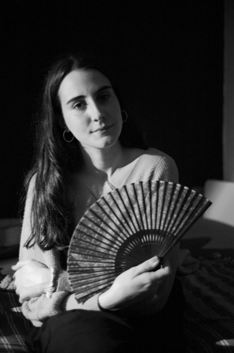 Woman with long hair and hoop earrings sitting on a bed holding an open folding fan