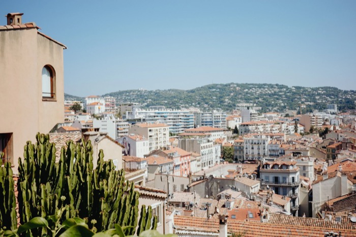 Terracotta rooftops of a Mediterranean town spreading toward wooded hills, tall cactus in the foreground