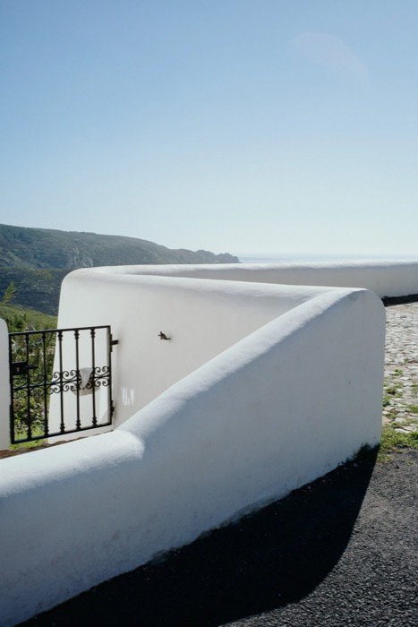 Curving whitewashed wall with a wrought-iron gate, coastline and sea in the distance