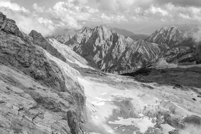 High alpine view over bare limestone slopes and snow patches to jagged peaks under piled clouds