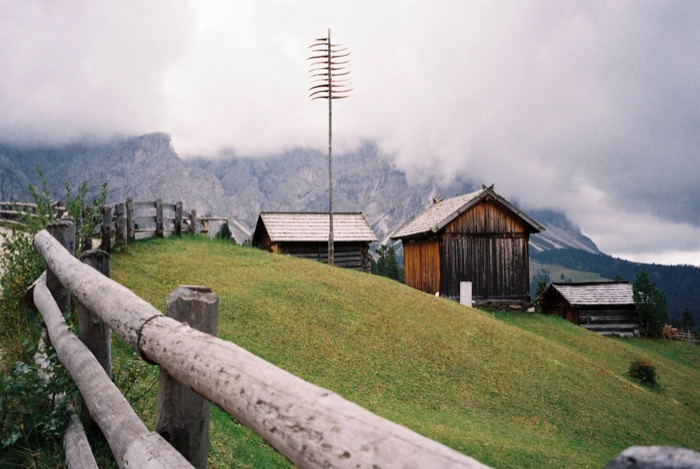 Wooden alpine barns on a grassy ridge below cloud-wrapped rock faces, split-rail fence in the foreground