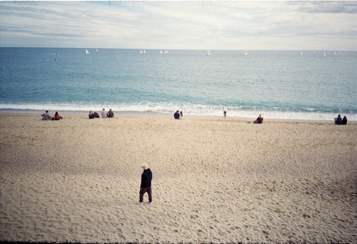 Man in black crossing a wide empty beach, small groups sitting at the waterline and sailboats offshore