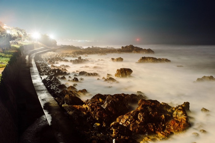 Long exposure of surf smoothed to mist over golden rocks beside a lit coastal path at night
