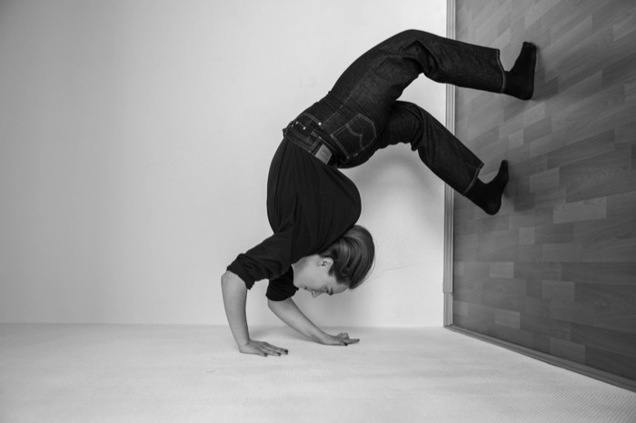 Woman balancing on her hands in a bare corner, legs bent up against the wall behind her
