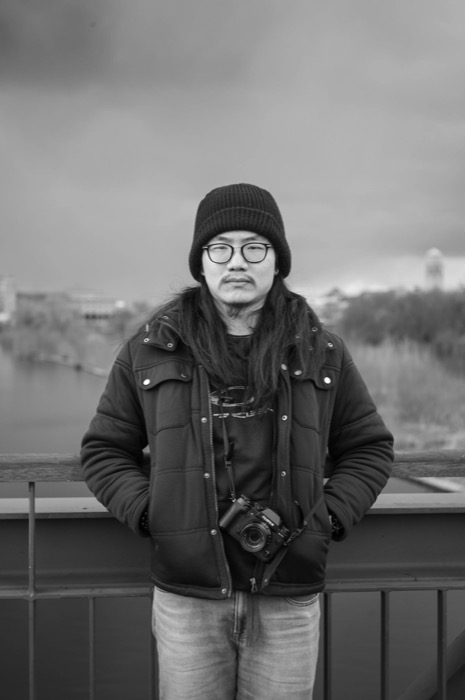 Long-haired man in beanie and padded jacket, camera round his neck, standing at a bridge railing over a river