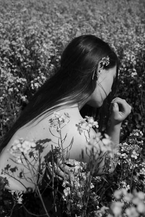 Woman with long hair and bare shoulders crouched in a flowering field, a blossom tucked behind her ear