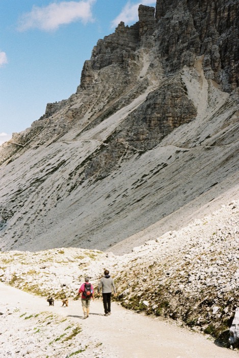 Two walkers with dogs on a gravel path beneath a vast pale scree slope and rock wall