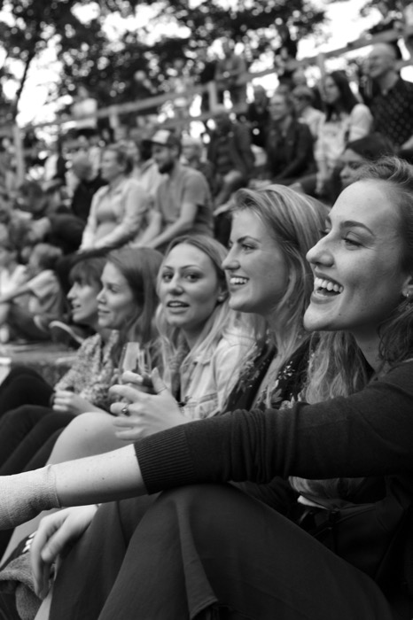 Row of young women laughing side by side on open-air stadium seating, a crowd rising behind them